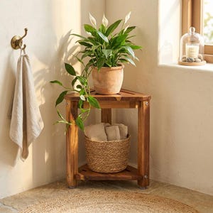 May include: A wooden corner shelf holds a potted plant with white flowers and green leaves. A woven basket with folded towels sits below. A beige towel hangs from a brass hook on the wall. A round jute rug is on the floor.