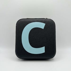 May include: A black, square case featuring a light blue letter "C". The case has a textured finish and a zip. The background is plain white.