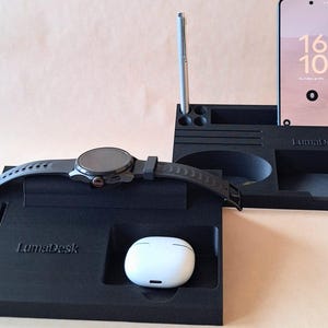 Puede incluir: Organizador de escritorio negro con el texto "LumaDesk" y un smartphone que muestra la hora 16:10. El organizador contiene un reloj inteligente, auriculares y un lápiz óptico. El diseño incluye varios compartimentos y ranuras.