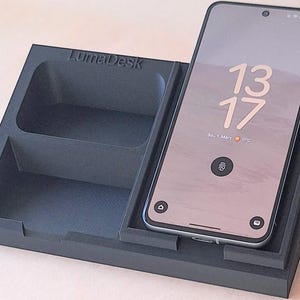 Könnte beinhalten: Ein schwarzer Schreibtisch-Organizer mit dem Text "LumaDesk". Auf der rechten Seite des Organizers wird ein Smartphone mit der Uhrzeit 13:17 angezeigt. Der Organizer hat ein Fach für das Telefon und einen separaten Ablagebereich.
