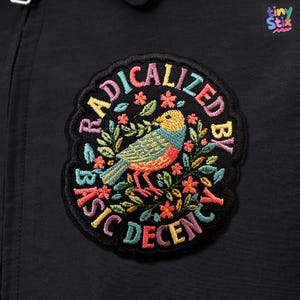 Peut inclure: &Eacute;cusson brod&eacute; avec le texte "RADICALIZED BY BASIC DECENCY" en lettres color&eacute;es. L'&eacute;cusson pr&eacute;sente un motif d'oiseau d&eacute;taill&eacute; avec des accents floraux dans les tons de jaune, bleu, orange et vert, sur fond noir.