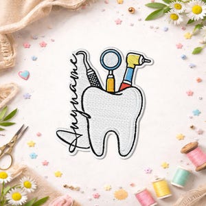 May include: An embroidered patch featuring a large white tooth with a black outline. The tooth is topped with a dental mirror, a scaler, and a dental drill in yellow, blue, and red. The word "Anyname" is written in black cursive script.