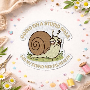Peut inclure: Un écusson brodé représentant un escargot de dessin animé avec une coquille brune et une expression triste, posé sur de l'herbe verte. Le texte indique "GOING ON A STUPID WALK" au-dessus et "FOR MY STUPID MENTAL HEALTH" en dessous en lettres dorées.