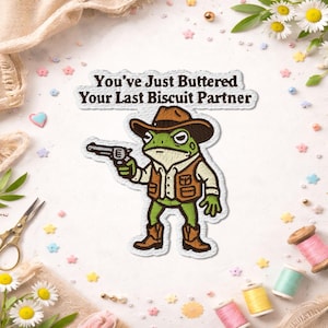 May include: Embroidered patch featuring a green frog in a cowboy hat and vest, holding a gun. The patch has the text "You've Just Buttered Your Last Biscuit Partner" in a white, outlined font. The frog is wearing brown boots.