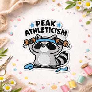 May include: An embroidered patch featuring a gray and white raccoon wearing a blue headband and lifting two brown dumbbells. The text PEAK ATHLETICISM is stitched in black above the raccoon, flanked by two light blue stars. A blue towel and water bottle are at the base.