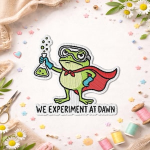 May include: An embroidered patch featuring a green frog wearing safety goggles and a red cape. The frog holds a glass flask filled with green liquid. Below the frog, the text reads WE EXPERIMENT AT DAWN in black capital letters.