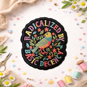 May include: Embroidered patch with a black border and the text "RADICALIZED BY BASIC DECENCY". The patch features a colorful bird surrounded by flowers and leaves. The text is in rainbow colors.