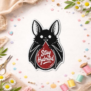 May include: An embroidered patch featuring a black bat with large ears and white eyes. The bat holds a large, red, blood-drop-shaped graphic in its wings. The text "Stay Hydrated" is written in white script across the center of the red drop.