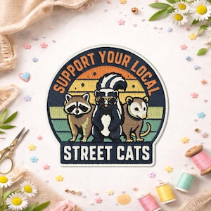 May include: Embroidered patch featuring a raccoon, skunk, and opossum with the text "Support Your Local Street Cats." The design includes a sunset background with a navy blue border. The skunk wears sunglasses.