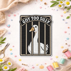 Peut inclure: Écusson brodé avec une oie blanche dans une cage noire, avec le texte "GOT TOO SILLY" au-dessus. Le patch a une bordure beige. Des fournitures de couture et des fleurs entourent le patch.