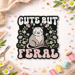 May include: Embroidered patch with a black background featuring a gray opossum illustration. The text "CUTE BUT FERAL" is embroidered in white and pink. Floral accents surround the opossum. Decorative sewing supplies are in the background.