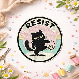 May include: Embroidered patch with the word "RESIST" in black lettering. A black cat with an angry expression kicks a gold crown. The patch has a pink, blue, and yellow background with a black border. Decorative elements surround the patch.