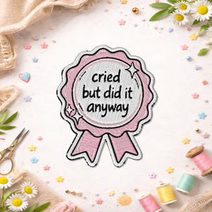 May include: A pink and white embroidered patch with the text "cried but did it anyway". The patch is in the shape of a ribbon with a scalloped edge and star accents. The background includes sewing supplies and floral accents.