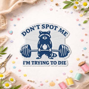 May include: Embroidered patch featuring a blue raccoon lifting a barbell. The text "DON'T SPOT ME" arches above the raccoon, with "I'M TRYING TO DIE" below. The patch is surrounded by decorative elements, including flowers and sewing supplies.