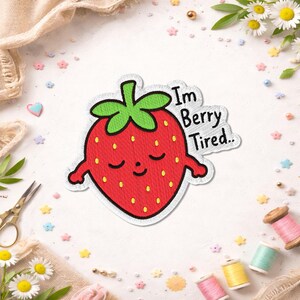 Peut inclure: Un écusson brodé représentant une fraise rouge avec une tige verte, des yeux fermés et un petit sourire. Le texte "Im Berry Tired.." est cousu en noir à côté de la fraise. L'écusson présente une bordure blanche et une finition en tissu texturé.