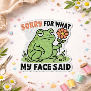 Peut inclure: Un écusson brodé représentant une grenouille verte assise sur de l'herbe, tenant une fleur orange. Le texte SORRY FOR WHAT MY FACE SAID est inscrit en lettres orange et noires. L'écusson a un fond blanc et une bordure découpée.