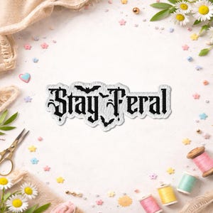 May include: A white embroidered patch with the words "Stay Feral" in a gothic font, featuring bat silhouettes. The patch is surrounded by decorative elements like flowers, spools of thread, and small star-shaped embellishments.