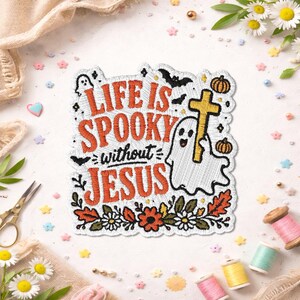 May include: An embroidered patch featuring the text "LIFE IS SPOOKY without JESUS" in orange and black thread. The design includes a white ghost holding a yellow cross, black bats, orange pumpkins, and a border of colorful autumn flowers and leaves.