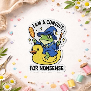 May include: Embroidered patch featuring a frog wizard in a blue robe and hat, riding a yellow rubber duck. The patch reads "I AM A CONDUIT FOR NONSENSE" in black text. The frog holds a wand and a corn dog.
