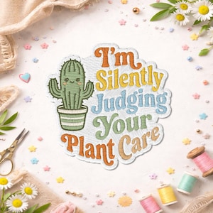 May include: An embroidered patch featuring a green cactus in a pot with a smiling face. The text reads I'm Silently Judging Your Plant Care in orange, yellow, blue, and green thread. The patch has a white background and a scalloped edge design.