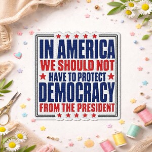 May include: A rectangular embroidered patch with a white background and a black border. The text reads IN AMERICA WE SHOULD NOT HAVE TO PROTECT DEMOCRACY FROM THE PRESIDENT. The words IN AMERICA, DEMOCRACY, and THE PRESIDENT are in blue.