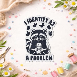 May include: An embroidered patch featuring a black and white raccoon holding a drink cup, surrounded by small butterflies. The text reads I IDENTIFY AS A PROBLEM in bold black letters. The patch has a white background and a die-cut border shape.