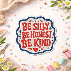 May include: A scalloped-edge embroidered patch featuring the red text BE SILLY BE HONEST BE KIND on a white background. The patch is outlined with a thick navy blue border and includes a small red heart icon below the text and navy blue stars.