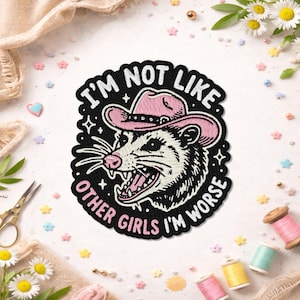 May include: Embroidered patch featuring a possum wearing a pink cowboy hat, with the text "I'm not like other girls I'm worse." The patch is black with white, pink, and black details. The patch is surrounded by sewing supplies and flowers.