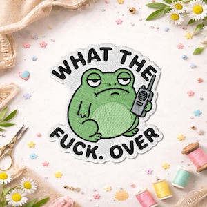 May include: Embroidered patch featuring a grumpy green frog holding a walkie-talkie. The patch has the text "WHAT THE FUCK. OVER" in black, surrounding the frog. The patch is on a white background with decorative elements.
