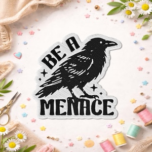 May include: Black embroidered patch featuring a raven with the text "BE A MENACE". The raven is detailed with white stitching, and the patch has a white border. The patch is surrounded by sewing supplies and flowers.