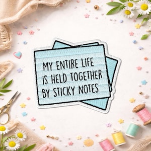 Peut inclure: Un autocollant bleu clair avec le texte "MY ENTIRE LIFE IS HELD TOGETHER BY STICKY NOTES" en noir. L'autocollant est entouré de fournitures d'artisanat, dont des bobines de fil, des ciseaux et des fleurs décoratives.
