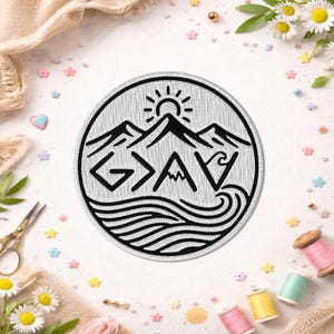 May include: A round, white embroidered patch with a black outline. The design features mountains, a sun, and waves, with the letters "GAV" in a stylized font. The patch is surrounded by sewing supplies and decorative elements.