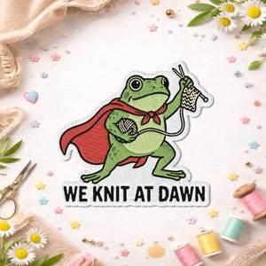 May include: Embroidered patch featuring a green frog wearing a red cape and holding knitting needles and yarn. The text "WE KNIT AT DAWN" is below the frog. The patch is surrounded by craft supplies and flowers.