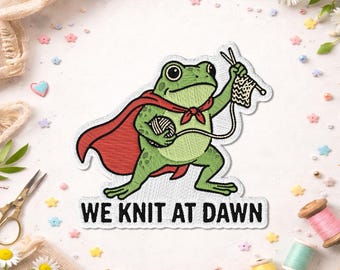 May include: Embroidered patch featuring a green frog wearing a red cape and holding knitting needles and yarn. The text "WE KNIT AT DAWN" is below the frog. The patch is surrounded by craft supplies and flowers.