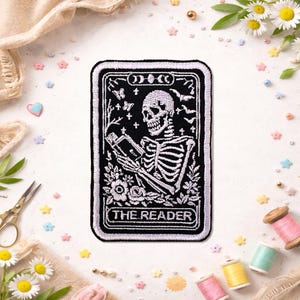 May include: Black and white embroidered patch featuring a skeleton reading a book, labeled "THE READER." The design includes moons, butterflies, and floral elements, with a black background and white border.