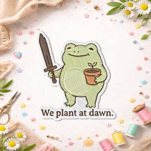 May include: An embroidered patch featuring a light green frog holding a dark brown sword in one hand and a small brown terracotta pot with a green sprout in the other. Below the frog, the text reads We plant at dawn in dark brown letters.