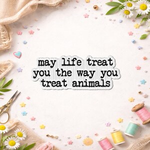 May include: A white sticker with the text "may life treat you the way you treat animals" in a typewriter font. The sticker is surrounded by daisies, sewing supplies, and decorative elements.
