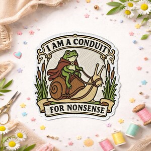 May include: An embroidered patch featuring a green frog wearing a brown cape and riding a snail. The design includes cattails on both sides and the text I AM A CONDUIT FOR NONSENSE in black lettering on a cream background with a decorative border.