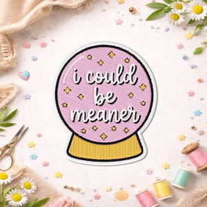 May include: A pink embroidered patch in the shape of a crystal ball with the text "I could be meaner" in white script. The patch has a black border and gold accents. The base of the crystal ball is gold.