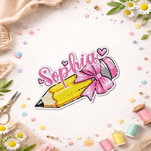 Peut inclure: Un écusson brodé représentant un crayon jaune avec une pointe noire et une gomme rose. Le prénom Sophia est brodé en lettres cursives roses au-dessus du crayon, agrémenté d'un grand nœud rose et de plusieurs petits cœurs roses autour.