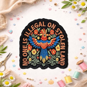 May include: Embroidered patch featuring a vibrant parrot design and the text "NO ONE IS ILLEGAL ON STOLEN LAND." The parrot is depicted in blue, orange, and yellow, set against floral embroidery on a black backdrop. The patch has a scalloped edge.