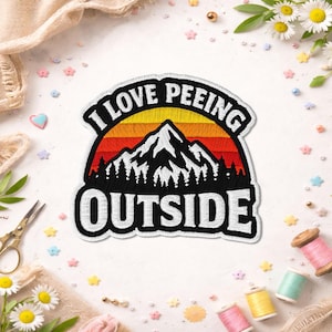 May include: Embroidered patch with the text "I LOVE PEEING OUTSIDE" above a mountain scene with a sunset background. The patch is black, white, red, orange, and yellow. The patch is surrounded by sewing supplies and decorative elements.