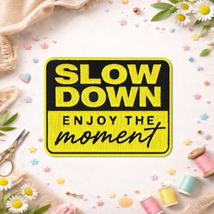 May include: A yellow and black embroidered patch with the words "SLOW DOWN" above "ENJOY THE moment". The patch is surrounded by sewing supplies, including spools of thread, scissors, and decorative flowers.