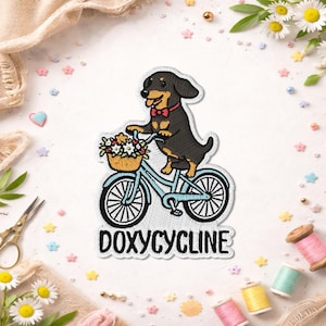 May include: Embroidered patch featuring a dachshund dog riding a light blue bicycle with a basket of flowers. The dog wears a red bow tie. The text "DOXYCYCLINE" is below the bicycle. The patch has a white border.