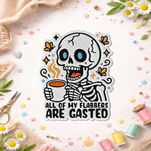 Peut inclure: Écusson brodé représentant un squelette aux yeux bleus tenant une tasse de café. L'écusson comprend des papillons et le texte "ALL OF MY FLABBERS ARE GASTED". Le design est noir et blanc.
