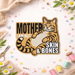 May include: Embroidered patch featuring a relaxed orange tabby cat with black stripes. The patch includes the text "MOTHER I AM BUT SKIN & BONES" in black and white. The patch is surrounded by sewing supplies and floral accents.