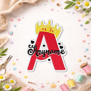 May include: An embroidered patch in the shape of the letter A, featuring a bright red fill with a white border. A yellow crown sits atop the letter, and the word Anyname is stitched in black cursive script across the center with small black hearts.