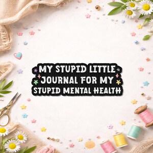 May include: Black patch with white text that reads "MY STUPID LITTLE JOURNAL FOR MY STUPID MENTAL HEALTH." The patch is surrounded by colorful stars. The background includes sewing supplies and decorative elements.