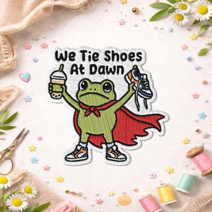 May include: An embroidered patch featuring a green frog wearing a red cape and trainers. The frog holds a boba tea in one hand and a trainer in the other. The text above the frog reads, We Tie Shoes At Dawn, in black embroidered lettering.