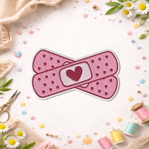 May include: Embroidered pink bandage patch with a red heart in the center. The bandage has a dotted pattern and is surrounded by a white border. The patch is designed for decorative use.